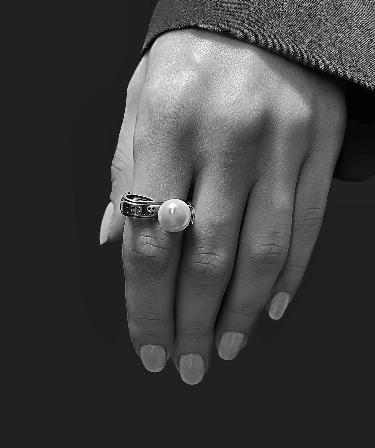 Mano en blanco y negro con un anillo de perla sobre fondo oscuro.