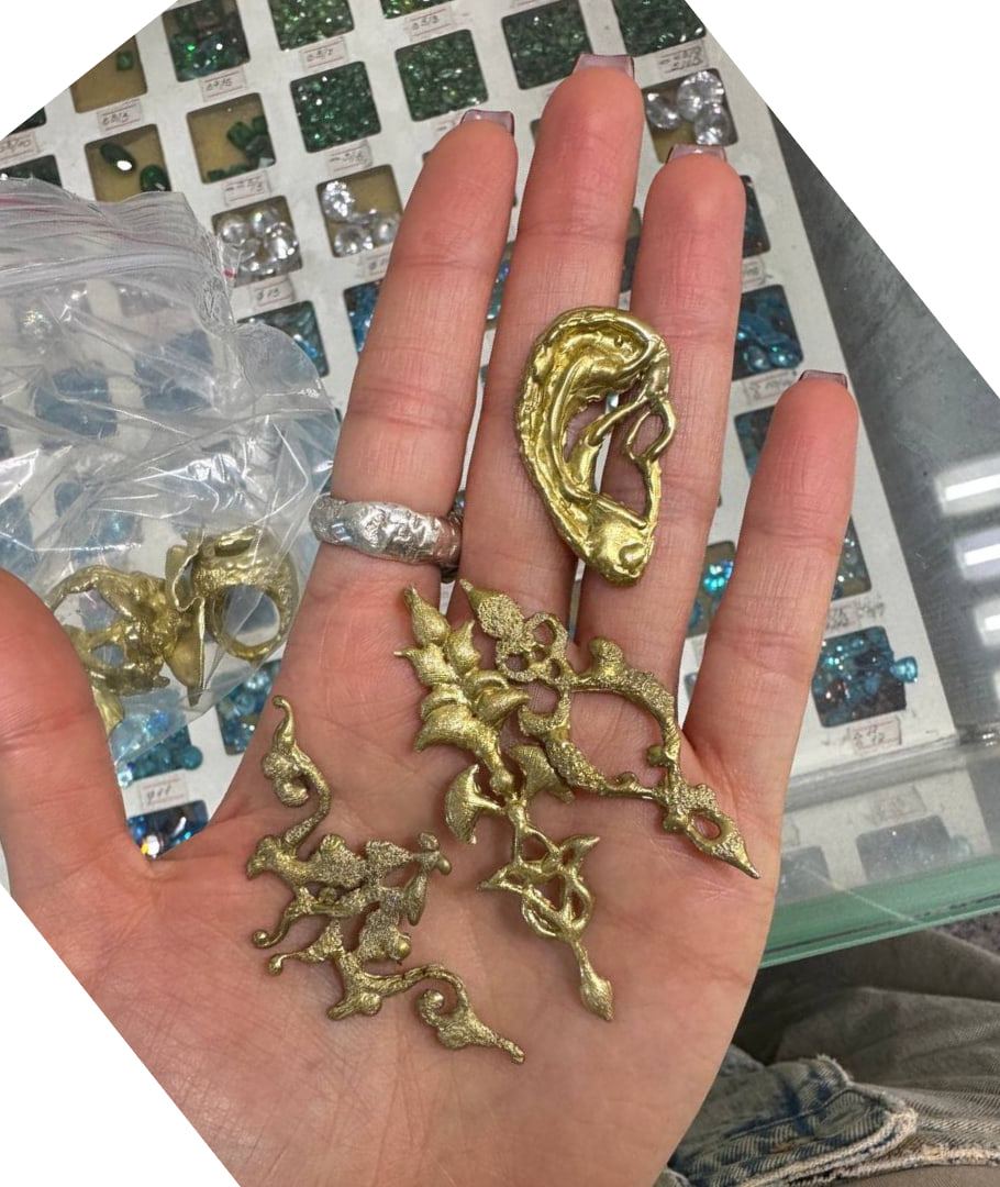 Piezas de ornamento recién fundidas, sin pulir, en la palma de una mano junto a una oreja de metal.