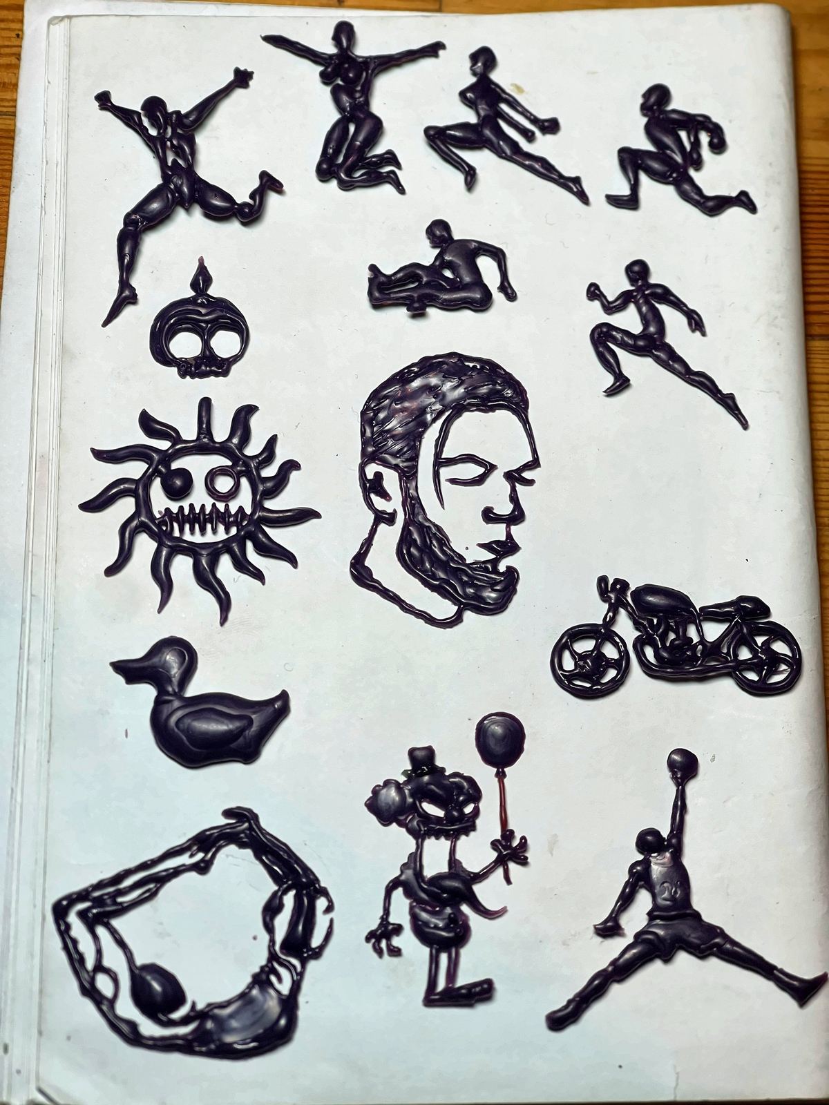 Figures of athletes, faces and a motorbike carved in wax on a white sheet.